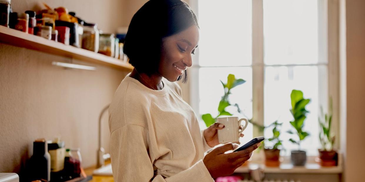 woman standing in the kitchen holding a cup of coffee and texting on her cell phone woman using a mobile phone at home