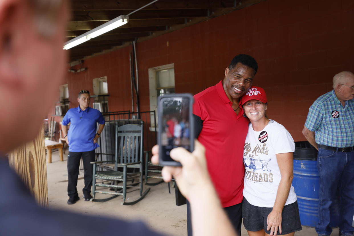Herschel Walker with his arm round a supporter in a baseball cap, as someone takes a picture with a cellphone.