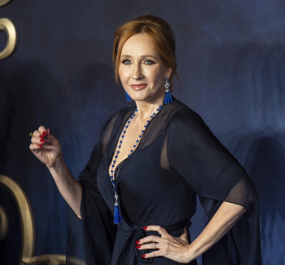 J.K. Rowling posing at an event, wearing a dark v-neck dress with blue necklace and earrings