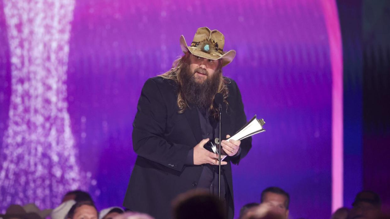 chris stapleton talks into a microphone on a stand as he stands on a stage, he looks to the left and wears a brown cowboy hat and black outfit