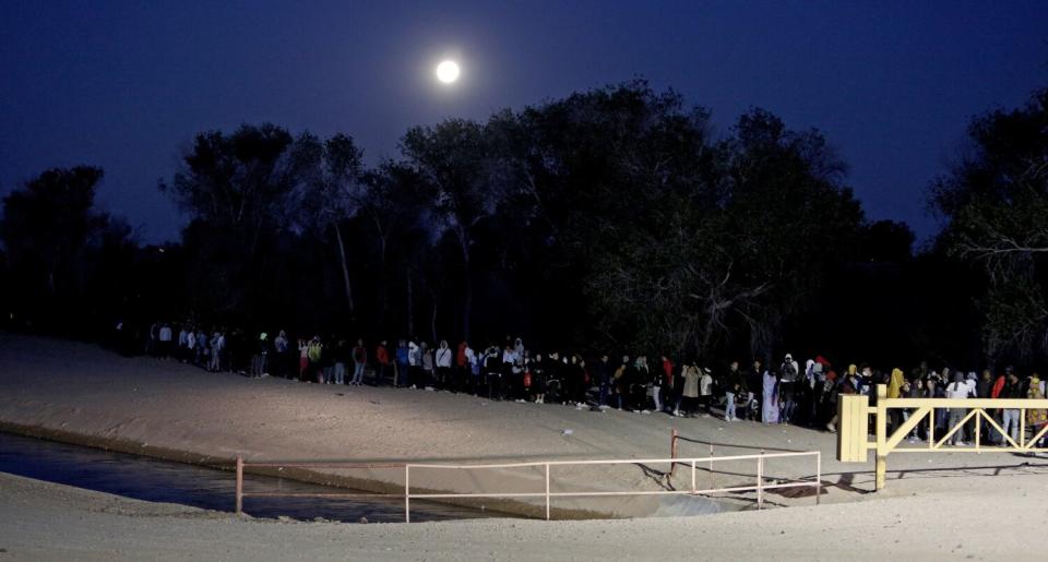 People forming a long line beside a grove of trees in a dusty field with a fence and gate, with the moon in a dark sky above