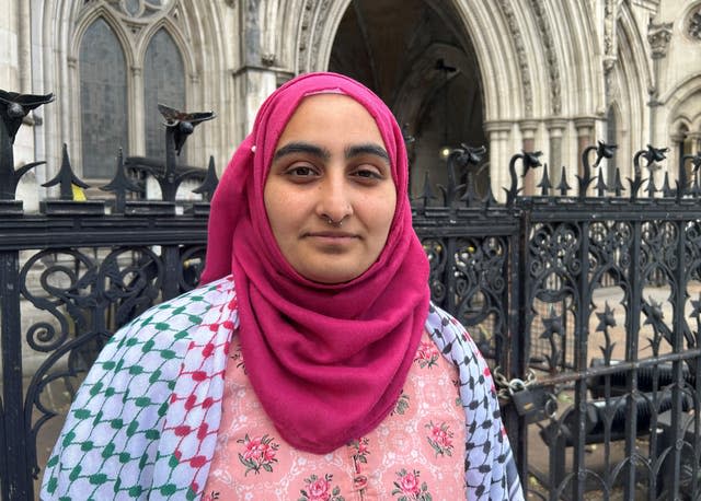 Close up a girl wearing a pink hijab, a pink flowered top and a white, pink and green scarf over her shoulders, standing in front of a stone building and black iron fencing