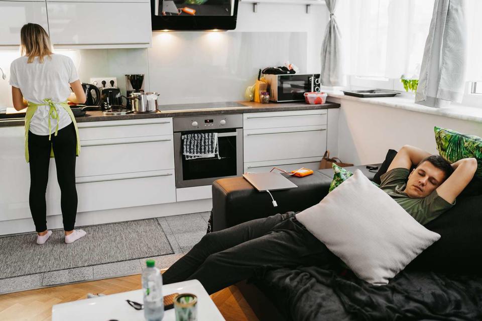 <p>Getty</p> Young woman preparing food in kitchen while man sleeping on sofa