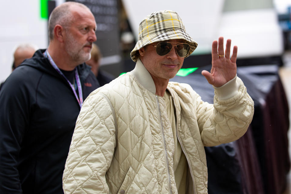 Brad Pitt waves to onlookers in the F1 paddock during practice ahead of the F1 Grand Prix in Burberry bucket hat, quilted bomber jacket, David Yurman ring, gold ring, David Yurman jewelry, 