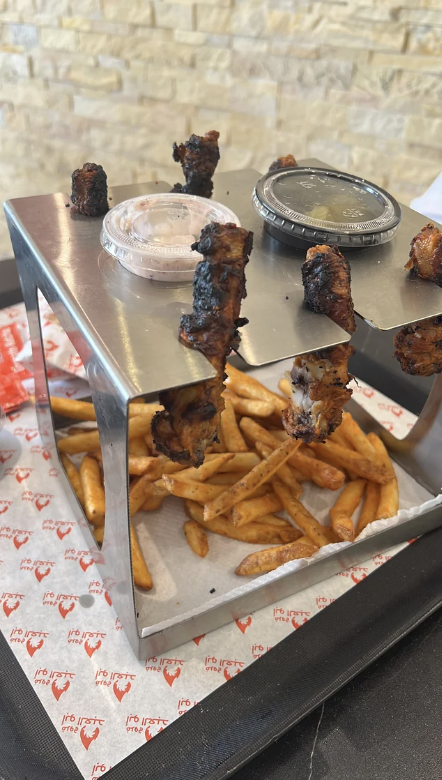 Skewers of grilled meat and dipping sauces placed on a metal rack over a pile of fries on a tray with restaurant branding