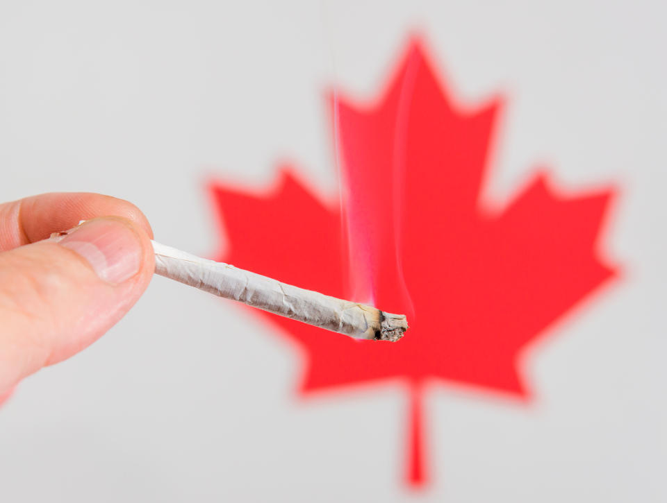 A lit cannabis joint being held in front of a red Canadian maple leaf.