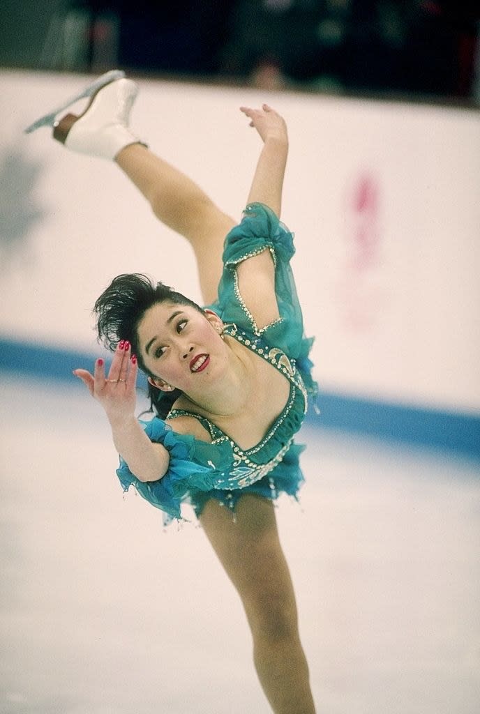 Kristi is wearing a ruffled skating dress as she skates on one leg