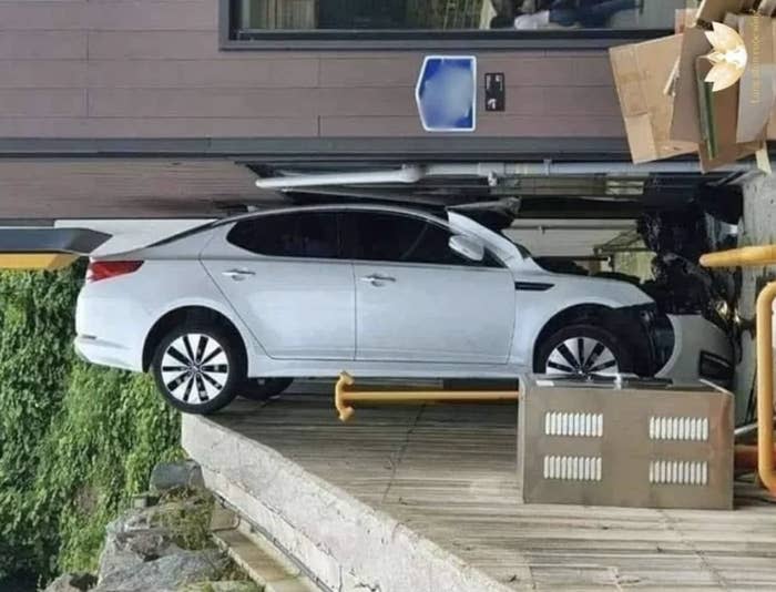 A car is parked on a steep ramp leading to an entrance. Boxes and equipment are stacked nearby. The car appears to have driven slightly over the edge.