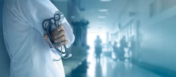 Medical professional in a white coat holding a stethoscope standing along the wall of a medical facility hallway with people out of focus in the distance.