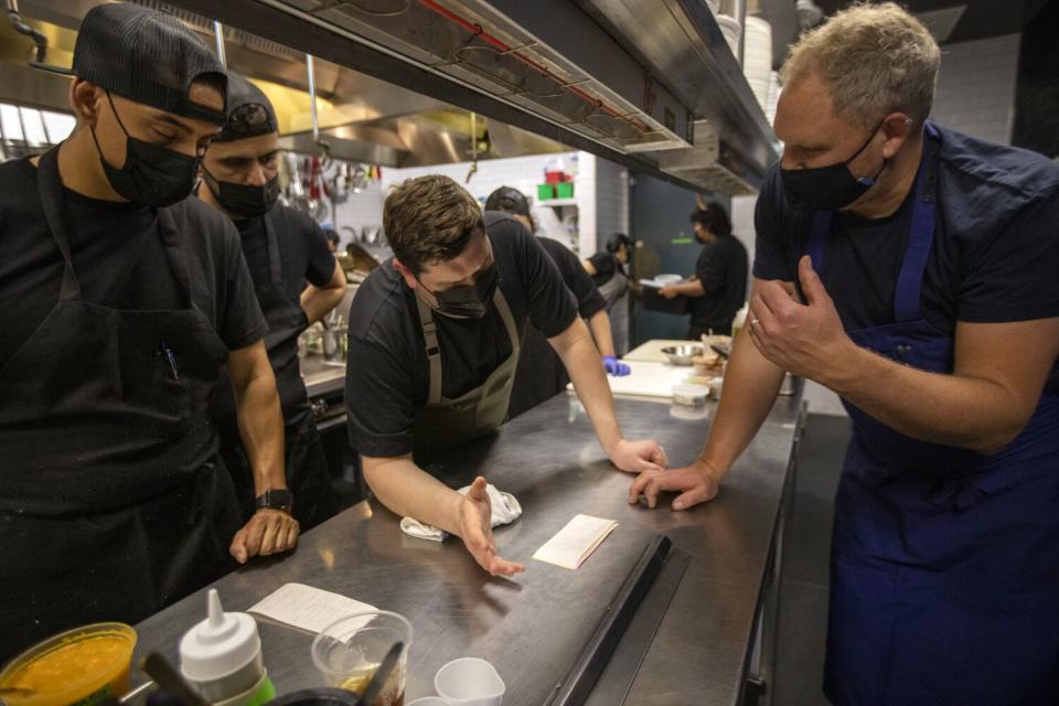 Several men in dark chef's clothing stand at a metal counter discussing a list.