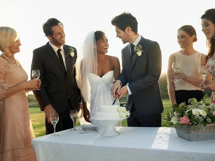 couple in wedding attire surrounded by guests and cutting into a cake
