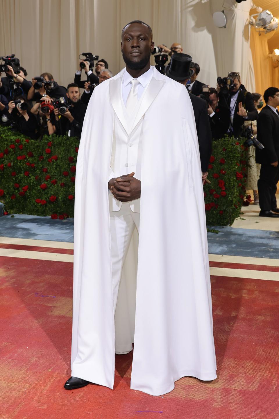 Stomzy in an all-white tuxedo with a floor-length white cape attached to the lapels of the jacket.