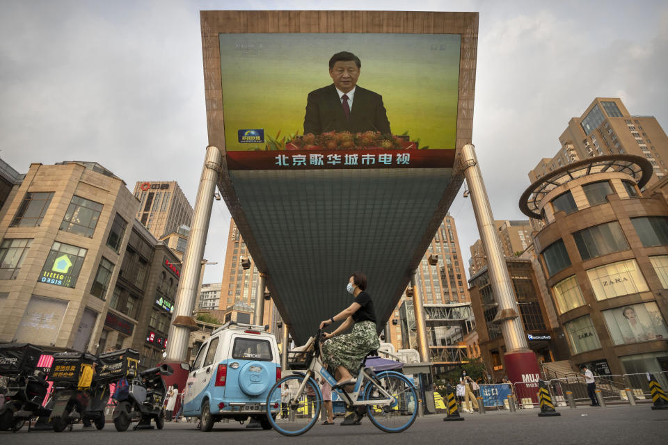 A woman wearing a face mask rides a bicycle past a large television screen at a shopping center displaying Chinese state television news coverage of Chinese President Xi Jinping's visit to Hong Kong in Beijing, Friday, July 1, 2022. (AP Photo/Mark Schiefelbein)