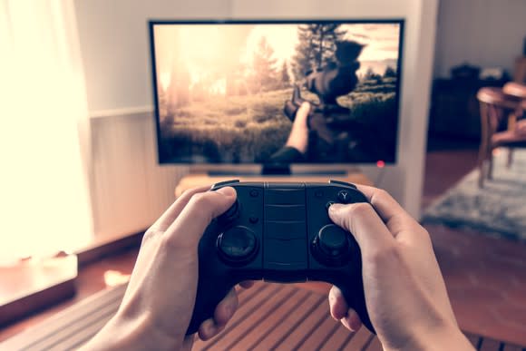 Hands holding a game controller with TV displaying a video game in the background.