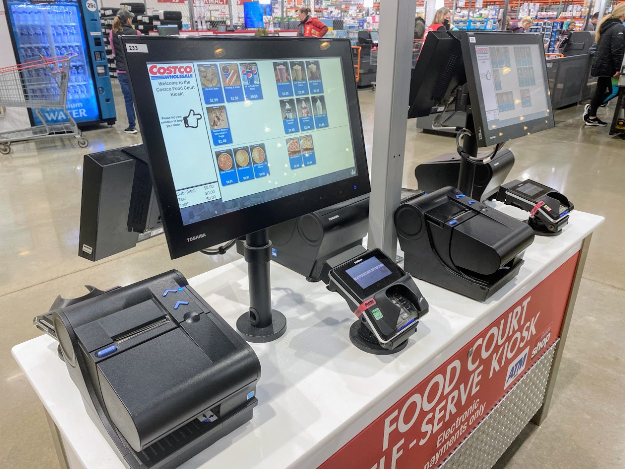 Costco food court self-serve ordering kiosk, ordering screen, separate card payment device, and separate receipt printer, cashier checkout lines and warehouse in the background