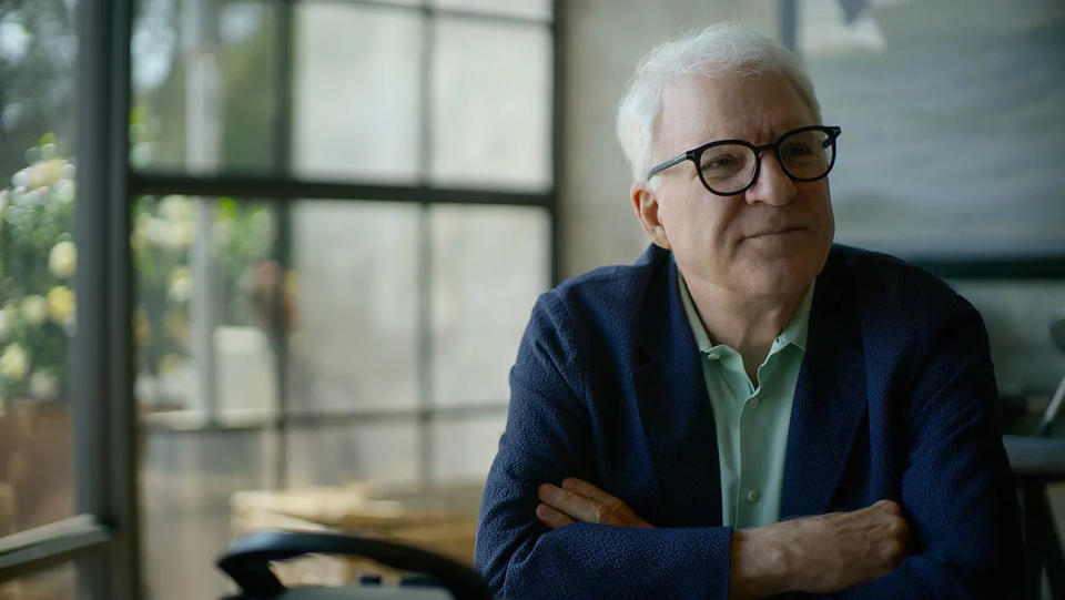 Steve Martin wearing glasses, sitting at a table in front of a large window