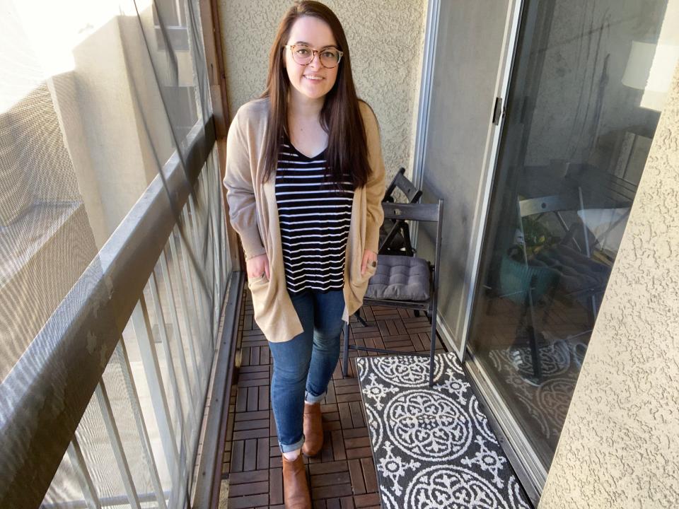 The writer wearing a Beige cadigan, black-and-white striped shirt, jeans, and brown boots on a balcony