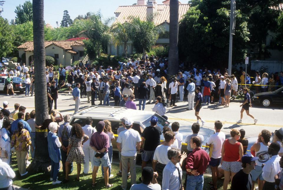 Dozens of people stand along a neighborhood street roped off with yellow tape near houses with terra-cotta roofs.