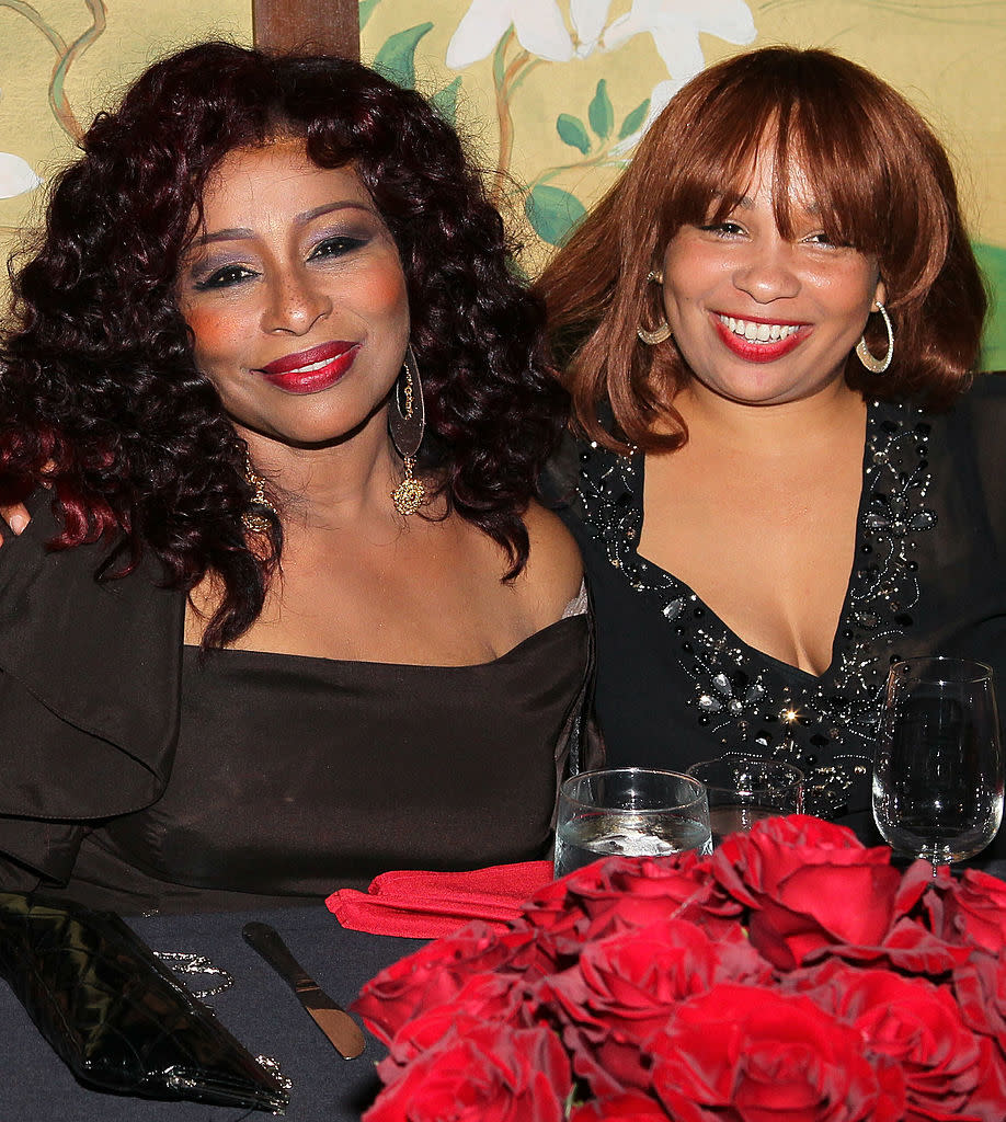 Chaka Khan and Indira Khan at an event, smiling and seated at a table with red floral centerpiece. Chaka is in an elegant one-shoulder dress