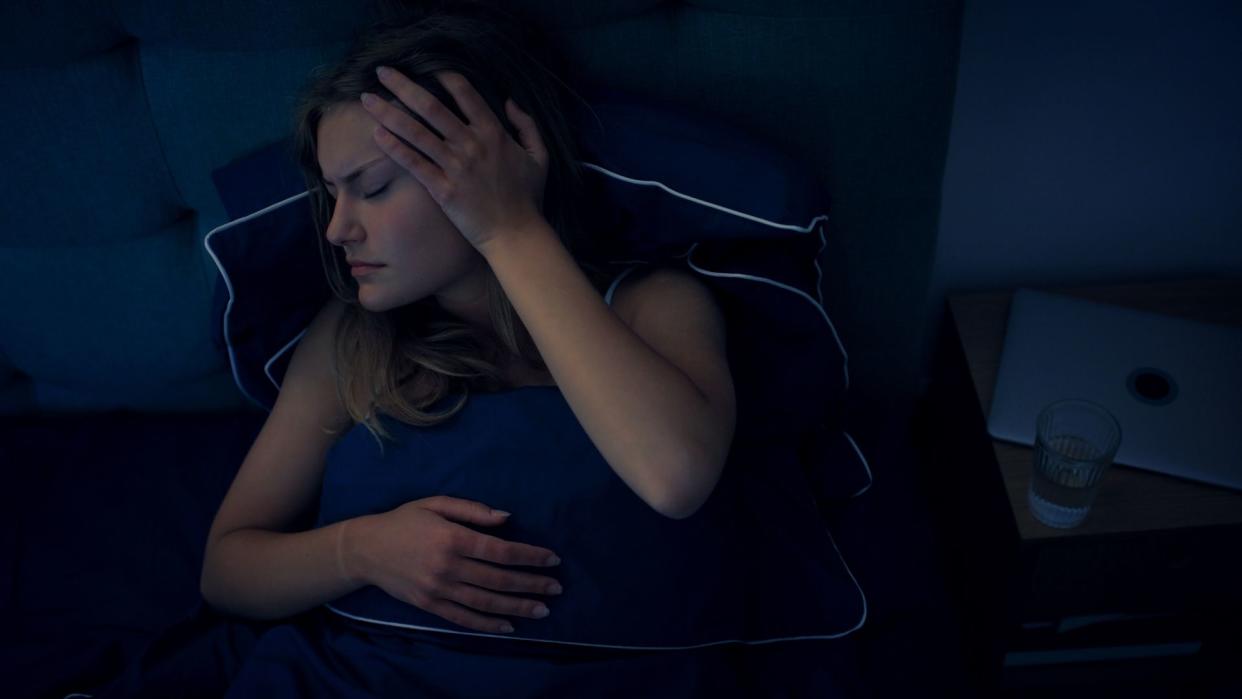  woman lying in bed in a dark room, holding a hand to her head as if in pain 