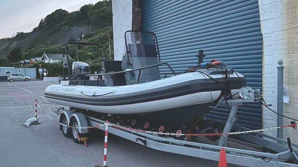 A grey electric powered inflatable boat moored up in front of metal shutters in a car park and fenced off with red and white chain