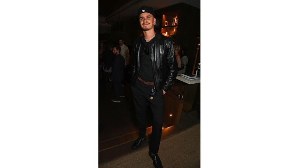 Romeo Beckham  in a black leather jacket, matching pants, and a black baseball cap stands confidently with a smile. His casual-chic look includes a chain necklace and key accessories.