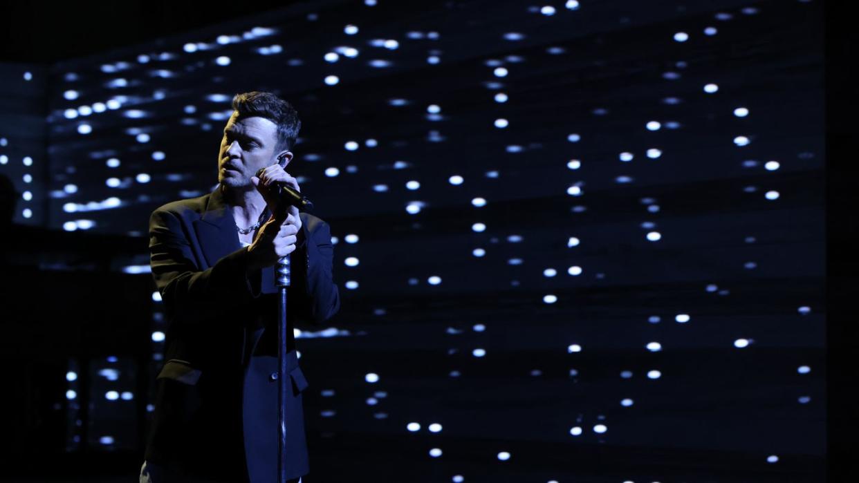 justin timberlake standing at a microphone in front of a dark white and blue lighted stage set