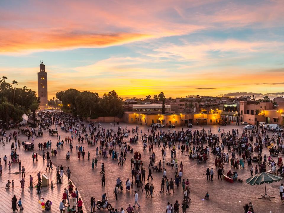 Hundreds of people walking around a city square in Morocco at sunset.