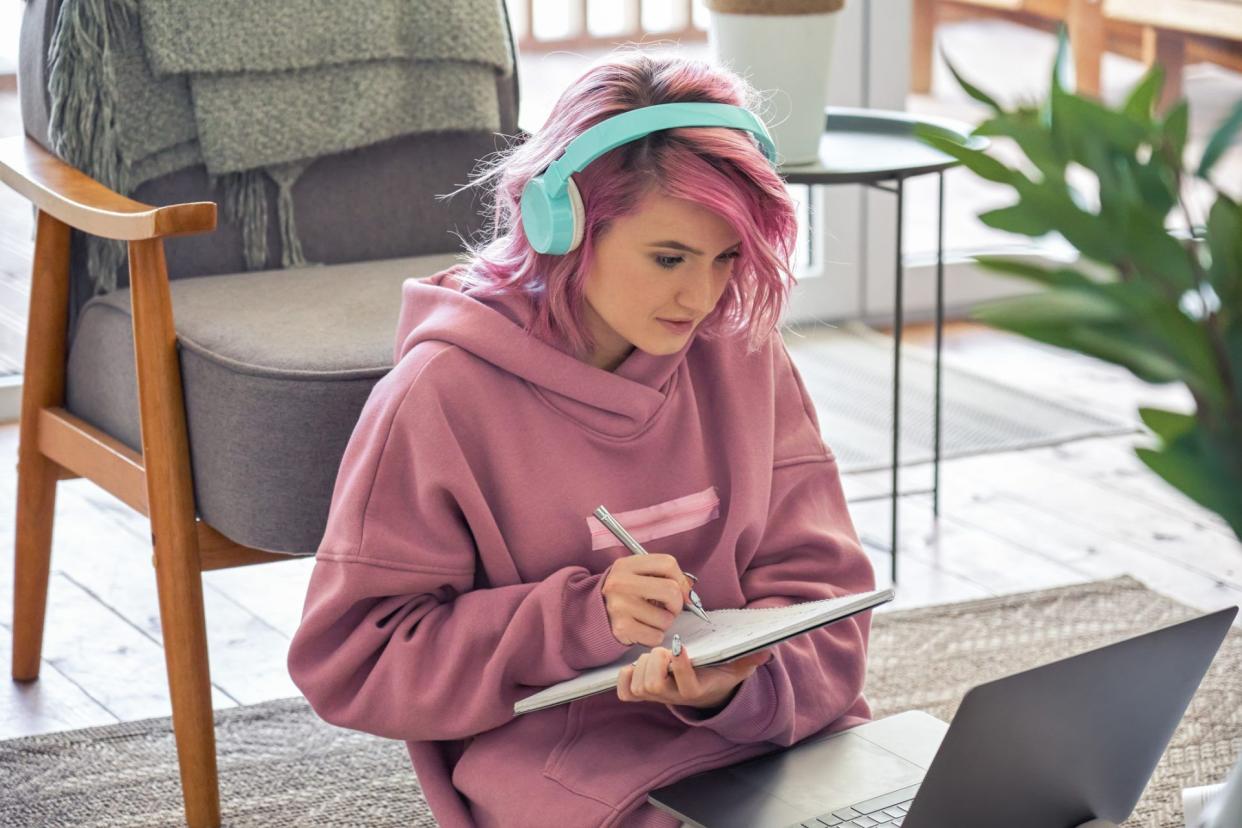 Focused hipster teen girl school college student pink hair wear headphones write notes watching webinar online video conference calling on laptop computer sit on floor working learning online at home.