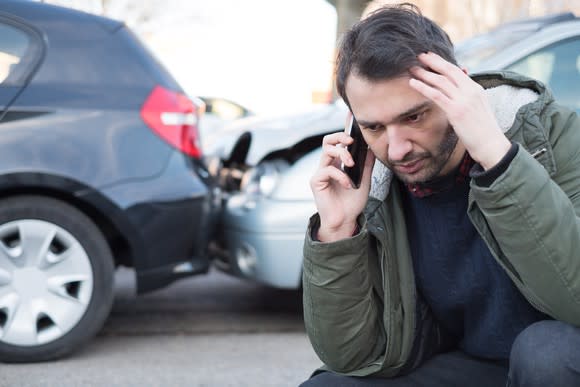 Man kneeling and talking on the phone in front of crashed cars.