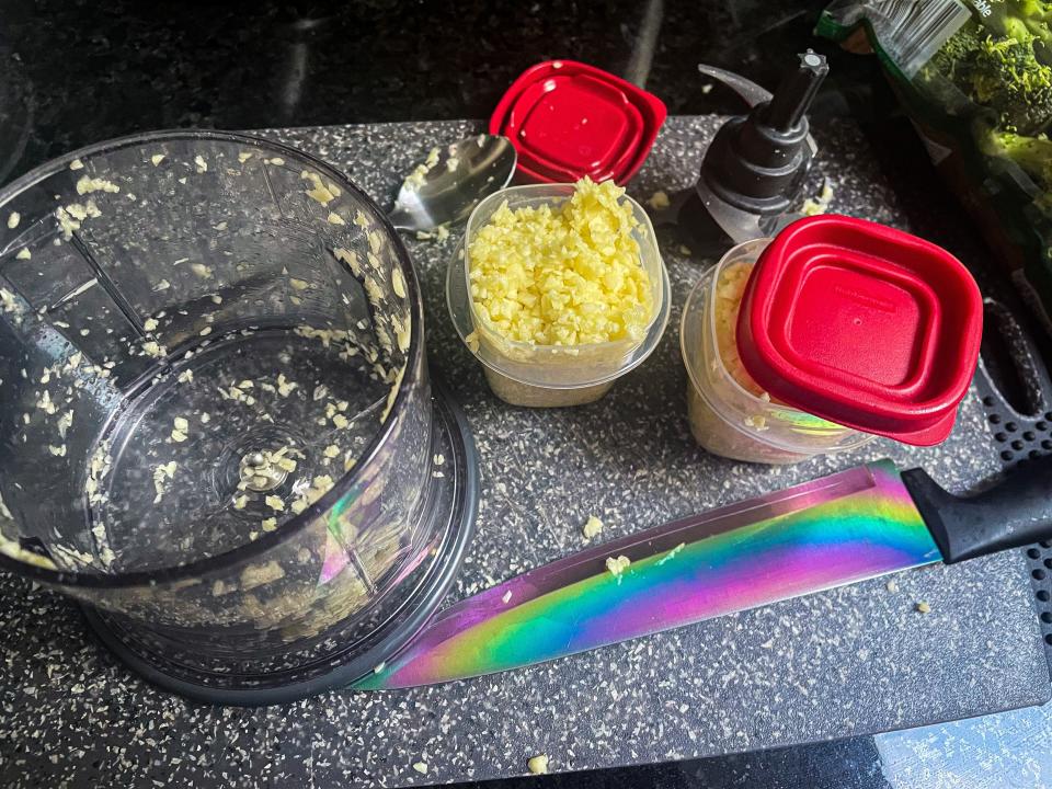 Food processor, kitchen knife with rainbow on the blade, and plastic cups with minced garlic sit on a counter