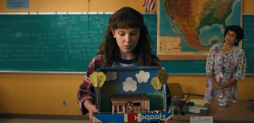 Eleven holding her diorama, wearing a baggy plaid shirt over a grey t-shirt