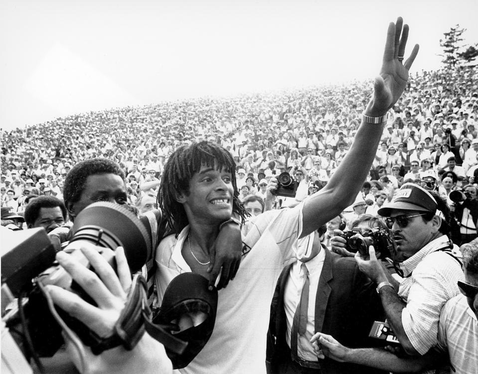 FILE - Yannick Noah, of France, waves to the crowd at Roland Garros stadium in Paris after defeating Sweden's Mats Wilander to win the French Open men's singles tennis title, June 5, 1983. Obscured at left hugging Yannick Noah is his father. . (AP Photo/Jacques Langevin, File)