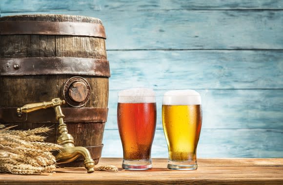 Two glasses of beer sitting next to a barrel and tap