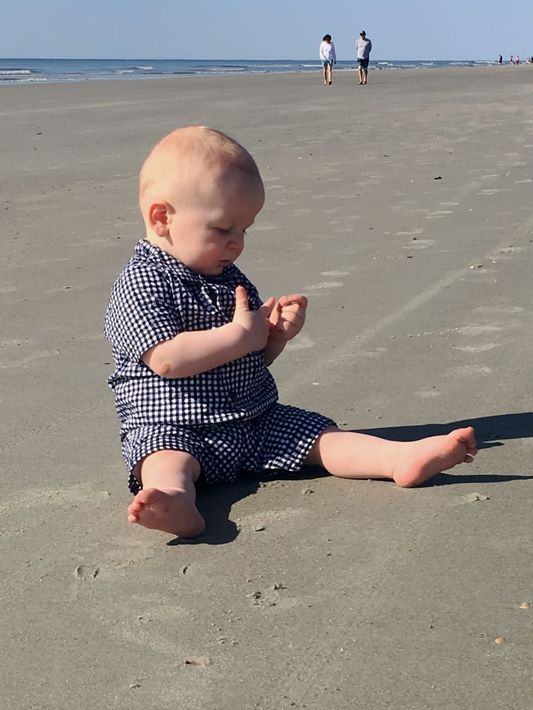 baby boy in blue and white plaid on the beach with people in the background