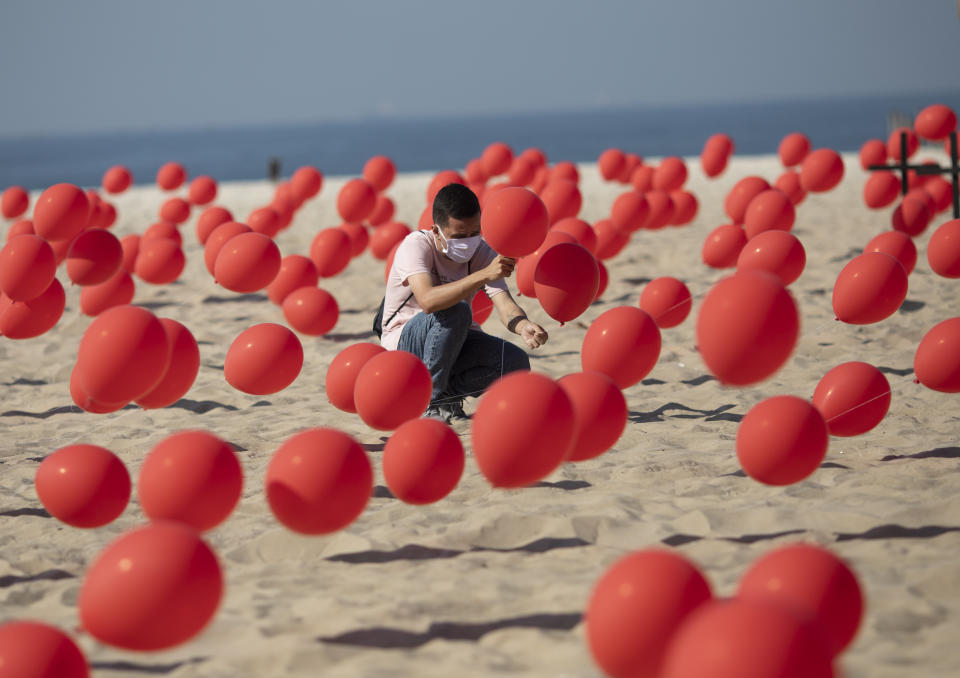 Un hombre ayuda a colocar globos rojos en la arena de la playa Copacabana durante un tributo organizado por Río de Paz para las víctimas de COVID-19 cuando el país se acerca a las 100.000 muertes por el nuevo coronavirus, en Río de Janeiro, Brasil, el sábado 8 de agosto de 2020. (AP Foto/Mario Lobao)