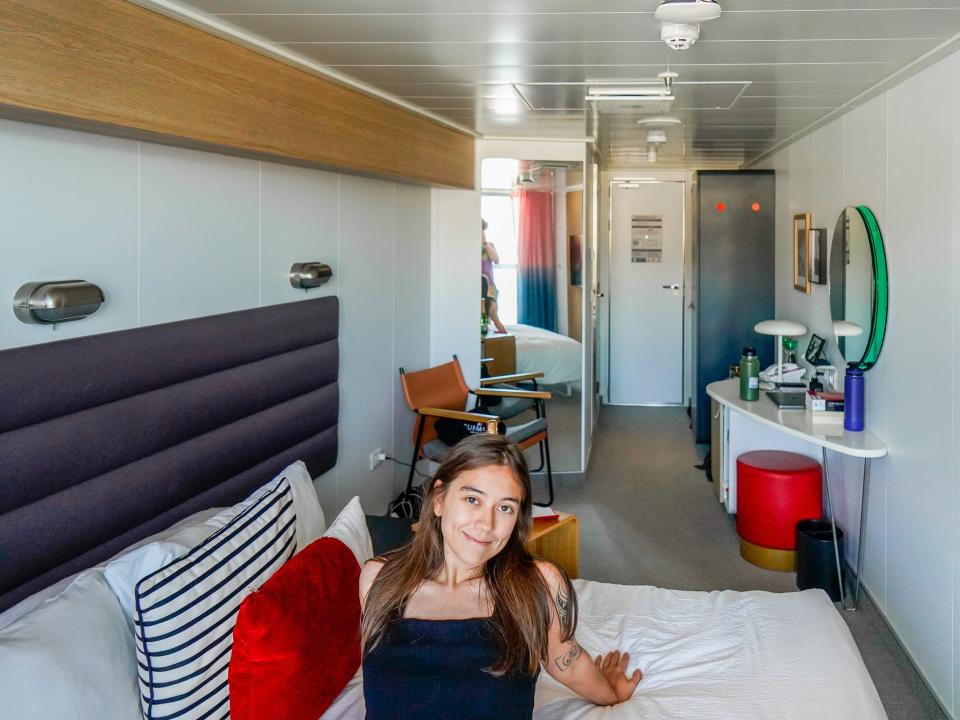 The author sits on a bed with white sheets and a red pillow on the left. Behind her is the cruise ship cabin