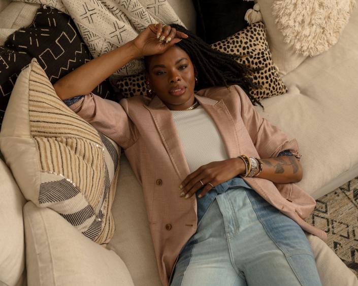 Interior designer Carmeon Hamilton poses on a sofa, wearing a dusty pink blazer over a white shirt with blue jeans.  She wears rose gold and silver jewelry, including rings, bracelets, and a watch.