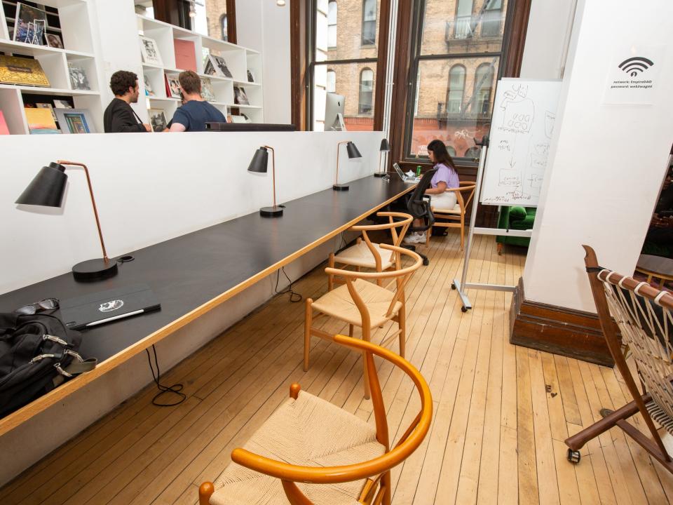 A long table with chairs, table lamps. Someone is working at the end of the table. People are standing nearby.