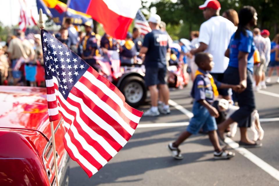 American flag flying from the back of a vintage red car in the foreground of a block party in a parking lot