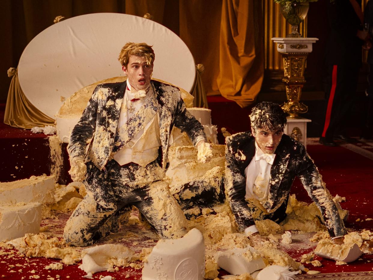 henry and alex kneel on a carpeted floor in red whit and royal blue, covered completely in smashed cake. they are dumbfounded