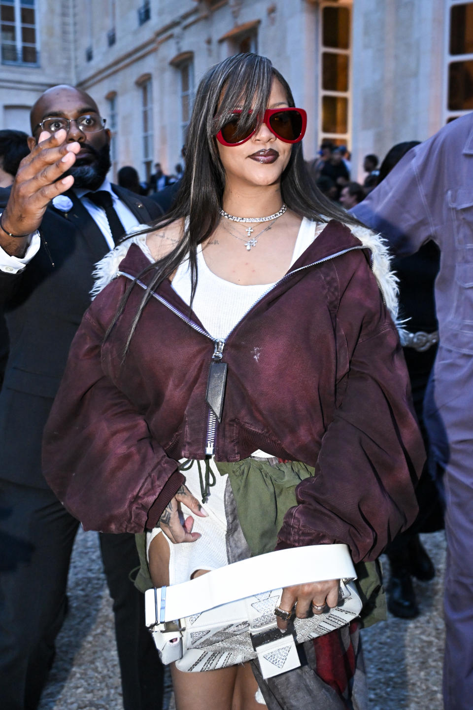 Rihanna is seen wearing oversized sunglasses, a layered outfit with a maroon jacket, and accessorized with necklaces, while walking outside an event