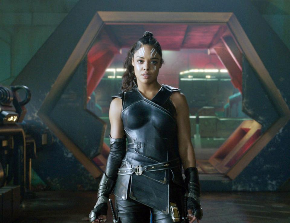 Tessa Thompson as Valkyrie in a sci-fi setting, wearing black leather armor with a fierce expression. She stands in a futuristic room with a mechanical background