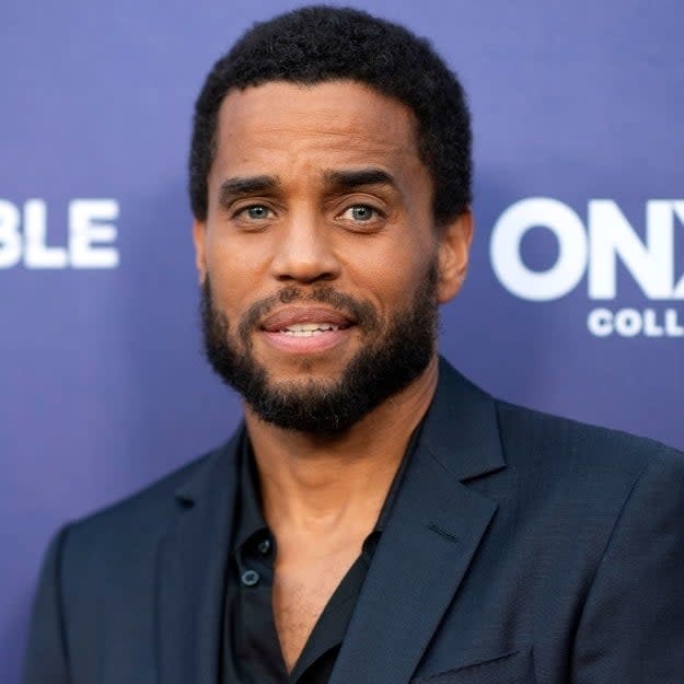 Michael Ealy at a celebrity event, wearing a dark suit with a black shirt. He smiles slightly, standing in front of a backdrop with the text 