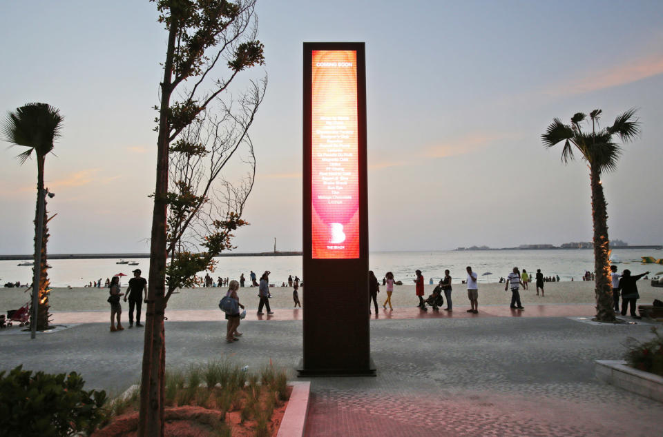 In this Saturday, April 19, 2014 photo, people walk past an advertising board with a camera installed in it and daily prayer time schedules at the beach in Dubai, United Arab Emirates. The ribbon of beach nestled along the towering skyscrapers rising from Dubai’s man-made marina district is a rare spot in the Middle East, bringing together the conservatively dressed and the bikini-clad in a multiethnic mix of those with means and those still striving for a more comfortable life. (AP Photo/Kamran Jebreili)