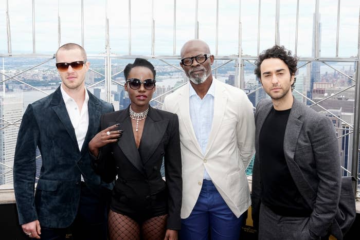 Lupita Nyong'o, Joseph Quinn, Djimon Hounsou, and Alex Wolff pose on a rooftop. Lupita wears a chic outfit with shorts, fishnet stockings, and statement sunglasses