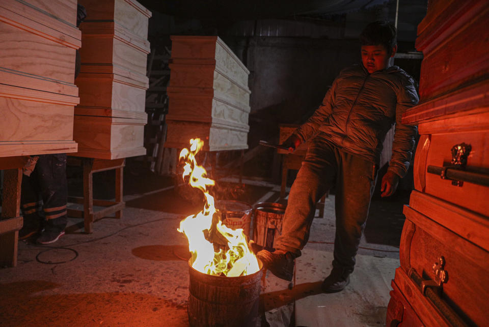 Cesar Ambrosio warms his feet on a fire while his father works in the finishing details of a production line of coffins at the Bergut Funeral Services' factory in Santiago, Chile, Thursday, June 18, 2020. The coffin production has had to increase up to 120%, according to Nicolas Bergerie, owner of the factory. His more basic coffin model is called the COVID model and is made to cope with the increase of deaths during the coronavirus pandemic. (AP Photo/Esteban Felix)