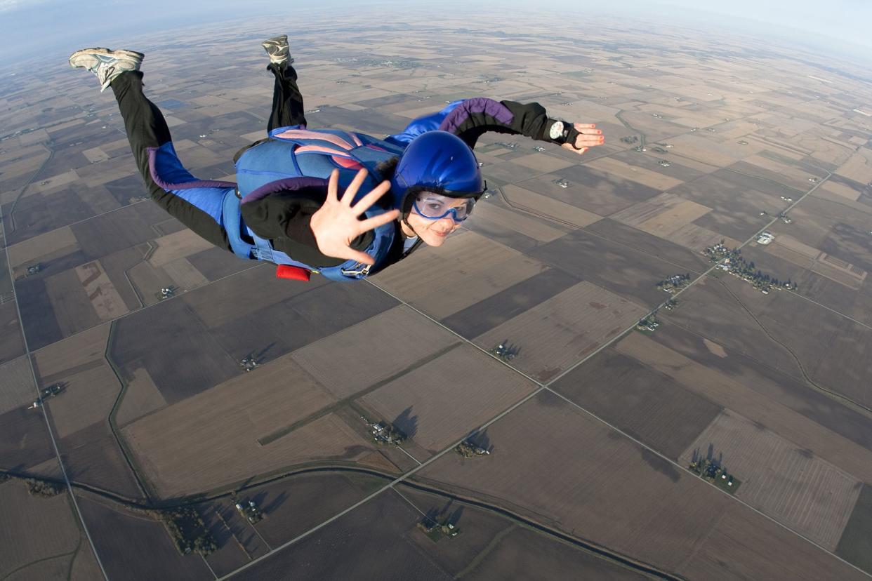 Young woman skydiving while mid-air smiling and waving to camera, over farm plots with a few roads, a curved horizon in the distance