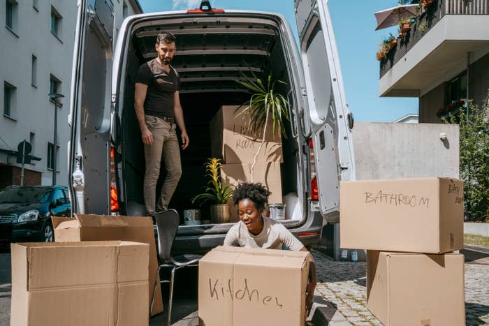 A man and a woman unload cardboard boxes from a moving van. The woman crouches while smiling, and the man stands inside the van
