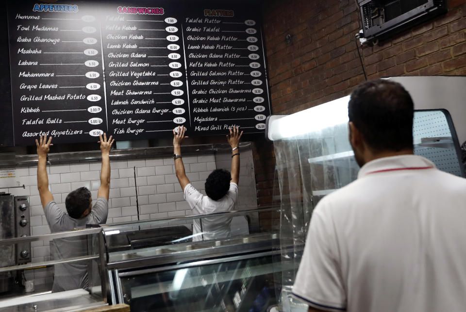 In this July 11, 2019, photo, Diaa Alhanoun, right, watches his business partner Mohammad Ayasrah, left, and son Nader, 18, hang a sign detailing their restaurant's extensive Mediterranean menu, in the Williamsburg neighborhood of Brooklyn in New York, days before the restaurant's opening. The menu features regional favorites. Everything on the menu is made by Alhanoun and Ayasrah. Desserts are prepared by Alhanoun's wife in the couple's home. (AP Photo/Kathy Willens)